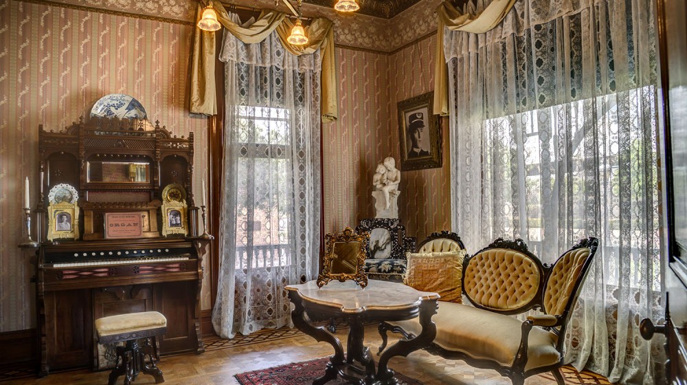 The front parlor at Rosson House Museum, with electric lighting, light pink wallpaper, a gold pressed tin ceiling, parquet wood floors, and ornate decorations.