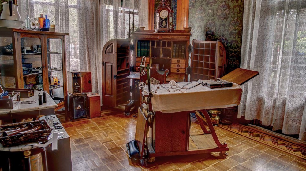 The doctor’s office at Rosson House, with dark green wallpaper, parquet wood flooring, a cabinet desk, an examining table and many other antique medical instruments.