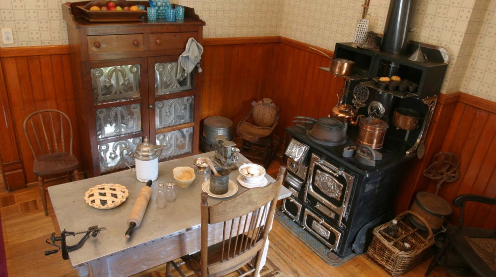 The kitchen at Rosson House with wood wainscotting and off-white wallpaper, parquet wood flooring, a wood burning stove, a pie safe, a kitchen table, and antique kitchen utensils and other implements.