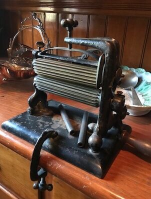 Rosson House pleating or crimping iron, used to press ruffles or pleats in clothing. It's made of cast iron, has two ridged rolls and a crank to turn them, and uses a clamp to attach it to a table to hold it still while running fabric through the rolls.