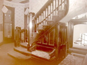 A picture of the entryway at Rosson House while the house was being restored. The wallpaper is light, the staircase and all other wood in the picture are very dark. The window under the stairs is broken, and there is dust everywhere.