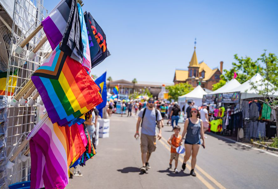 A picture of the Rainbows Festival at Heritage Square, with festival tents and booths lining the street, people enjoying themselves, beautiful and colorful pride flags, and Rosson House in the background.