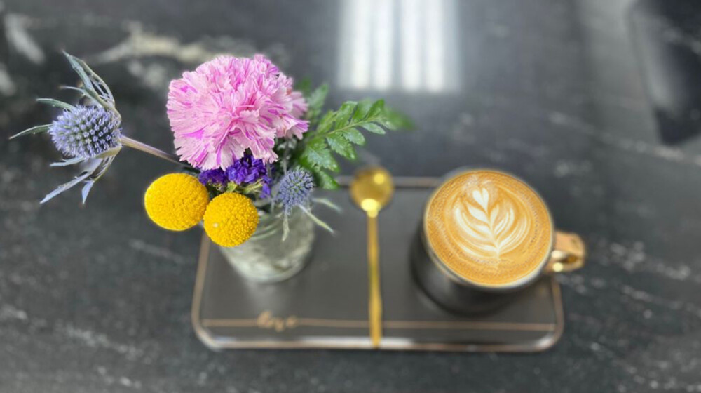 A picture of a tasty looking cup of coffee with a heart shaped latte art in the foam on top, next to bright and beautiful looking flowers.