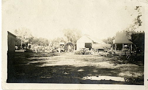 A black and white picture of Alejandro and Maria Silva's ranch, circa 1920. The picture shows a few vehicles with large, spoked wheels that would have been pulled by horses or other livestock. It also shows several ranch buildings, trees, and what appears to be a large water wheel.