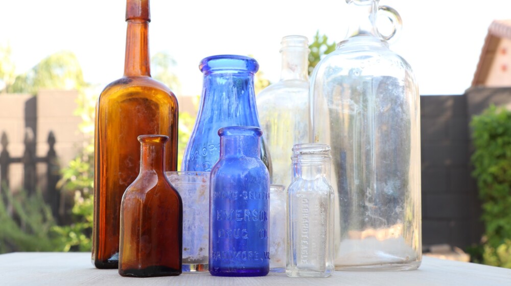 A picture of nine bottles or glass containers, all of different shapes and sizes, from the Heritage Square museum collection. There are two brown bottles, two cobalt blue bottles, and the remainder are clear glass.