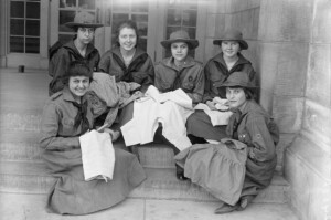 A black and white picture, taken in 1918, of six Girl Scouts in their uniforms, sitting on steps and sewing.