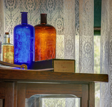 A picture of Victorian Era medicine bottles on display in the Rosson House Museum doctor's office. On the right is a large, dark brown bottle with "William Radam’s Microbe Killer" molded directly in the glass. On the left is a small, clear bottle with a label for Gombault's Caustic Balsam.