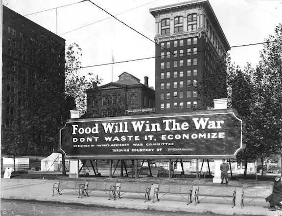 A black and white picture of a building with a banner out front that reads, "Food Will Win the War - Don't waste it, economize". Circa 1918.