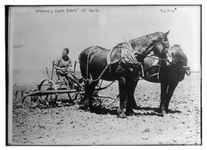 A black and white photograph of a woman doing work for the Women's Land Army during the first World War. She is sitting on a piece of farm equipment that is being pulled by two large horses.