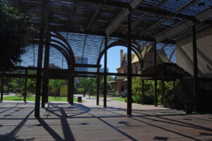 A picture from inside the Lath Pavilion at Heritage Square, looking out at the Rosson House Museum and Visitor Center. Verdant green grass and trees and a perfect, cloudless blue sky can be seen beyond.