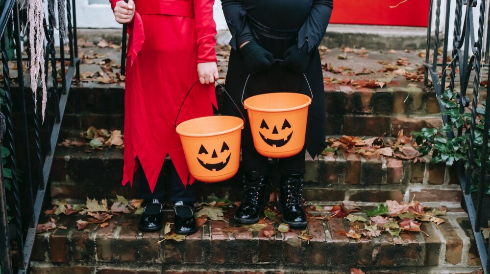 A picture of two children, standing on steps in front of a building and dressed up for Halloween with plastic jack o' lantern buckets.