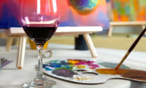 A picture of a canvas being painted, with paints and a paintbrush, and a glass of wine in the foreground.