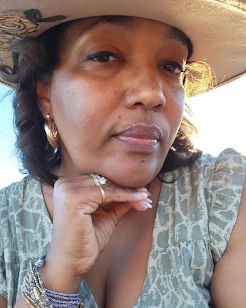 A picture of a woman wearing a hat, with her chin resting on her hand, looking at the camera.