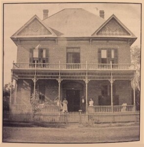 A black and white picture of a Victorian Era house with two levels and a wide front porch.