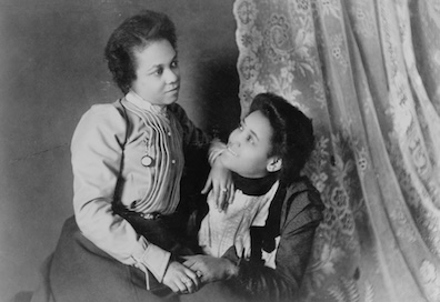 A picture of two Black women, sitting closely to each other and holding hands, taken in 1900 by W E B DuBois.