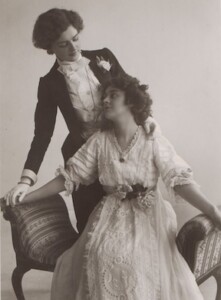 A black and white photograph, taken in 1907, of a woman wearing a light-colored dress, sitting in a chair, with another woman standing behind her, wearing a tux.