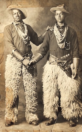 A antique photograph in sepia tones of two Black cowboys holding hands.