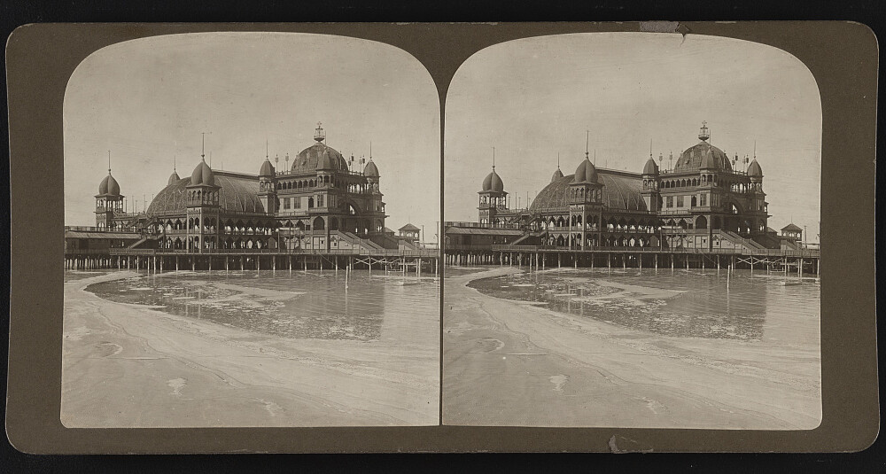 An old, black and white stereoscope card, with two pictures side by side of the Saltair pavilion in 1906. It stands three stories above the lake with rounded turrets and open air spaces.