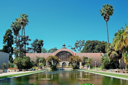 A picture of the Balboa Park Botanical Building, a large wood lath structure under a blue sky with a pond and palm trees in front.