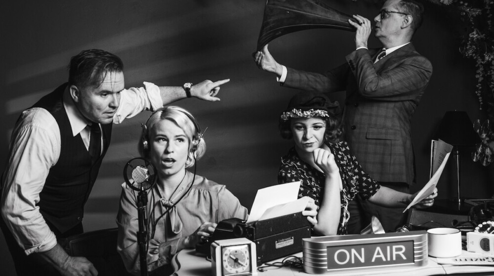 A present day black and white photo of people acting during a stage play of War of the Worlds, showing a radio program desk and four people.