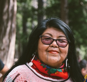 A photograph of the a smiling indigenous woman, with long dark hair, wearing a red scarf and glasses with black frames, and with a forest in the background.